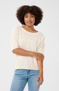 Pull en tricot texturé de couleur crème clair avec un large col et des manches courtes, associé à un jean bleu. Design simple, sans motifs ni accents.