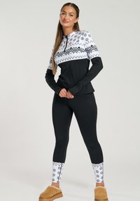 Set atletico nero con una parte superiore bianca a fantasia con design geometrici, leggings neri slim-fit e comode scarpe slip-on marroni.
