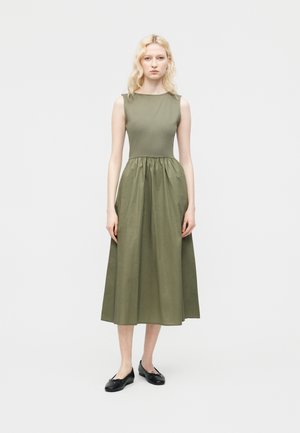 Γυναίκα με ανοιχτό ξανθά μαλλιά φοράει αμάνικο μεσαίο φόρεμα σε ελιά πράσινο χρώμα και μαύρες μπαλαρίνες, μπροστά σε απλό λευκό φόντο.