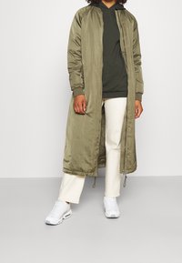 Długa kurtka bomberka w kolorze oliwkowej zieleni z zamkiem z przodu, ściągaczami na rękawach i bocznymi kieszeniami, nałożona na ciemnozieloną bluzę z kapturem i kremowe spodnie.