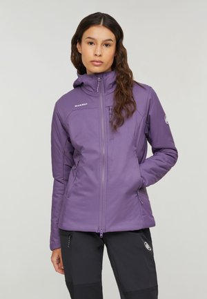 Giovane donna con lunghi capelli mossi, che indossa una giacca Mammut viola e pantaloni neri, in piedi con una mano in tasca davanti a uno sfondo semplice.