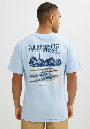 Hombre que lleva una camiseta azul claro con un gráfico de una regata de remo, la Torre Eiffel y árboles, de pie de espaldas sobre un fondo blanco.