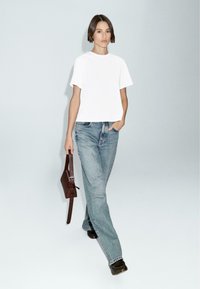 T-shirt bianco a maniche corte abbinato a jeans a vita alta blu chiaro. Borsa in pelle marrone scuro tenuta nella mano sinistra. Scarpe nere.
