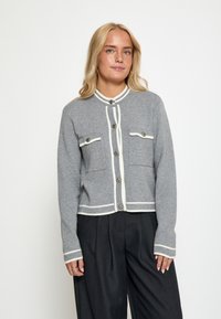 Šedý pletený cardigan s bílými obrysy, má dvě náprsní kapsy, zaoblený límec a zapínání na knoflíky. Měkká textura a přiléhavý střih.