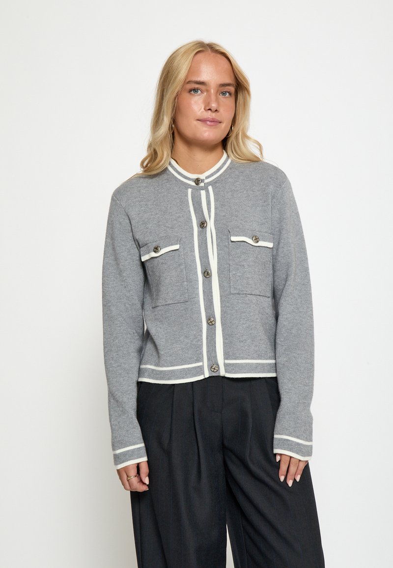 Šedý pletený cardigan s bílými obrysy, má dvě náprsní kapsy, zaoblený límec a zapínání na knoflíky. Měkká textura a přiléhavý střih.