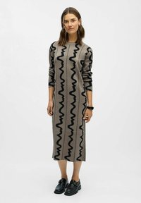 Bežinė megzta suknelė su juodais bangų raštais, ilgomis rankovėmis, laisvu siluetu, iki kelių, derinama su juodais batais, paprastas dizainas.