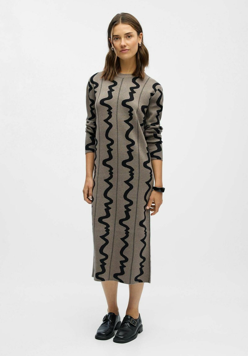 Bežinė megzta suknelė su juodais bangų raštais, ilgomis rankovėmis, laisvu siluetu, iki kelių, derinama su juodais batais, paprastas dizainas.