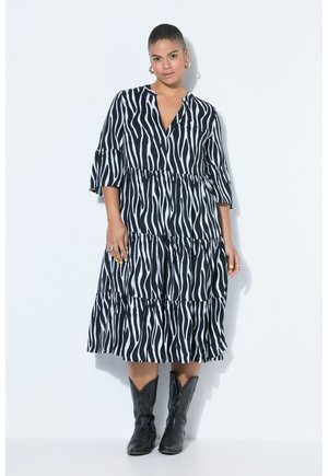 Kobieta w czarno-białej sukience midi w wzór zebry z rękawami 3/4 i czarnych kowbojskich butach, stojąca na białym tle.