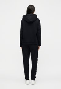 Άτομο που στέκεται γυρισμένο προς τα πίσω, φορώντας μαύρο φούτερ με κουκούλα και αντίστοιχο παντελόνι με λευκό κείμενο στα πλαϊνά ραφές και λευκά αθλητικά παπούτσια.