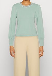 Maglione a maniche lunghe verde menta chiaro con scollatura rotonda decorato con accenti di perline, abbinato a pantaloni beige a gamba larga.