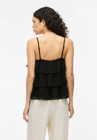 Canotta nera a più strati con sottili spalline regolabili, caratterizzata da volant a strati e una texture semi-trasparente, abbinata a pantaloni di colore chiaro.