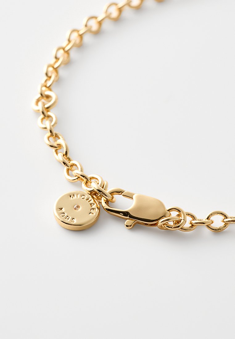 La pulsera de cadena de metal dorado presenta un cierre de langosta y un charm redondo con texto grabado. Los eslabones son ovalados y pulidos.