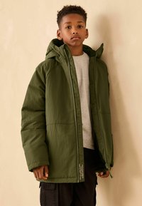 Grüne Pufferjacke mit Kapuze, ausgestattet mit einem Frontreißverschluss, zwei Seitentaschen und gerippten Manschetten, getragen über einem grauen Shirt. Schlichte, strukturierte Stoffqualität.