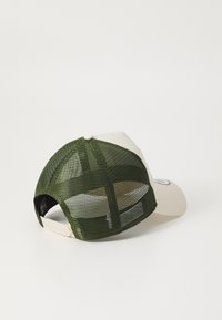 Gorra de béisbol con la parte frontal color crema y la parte trasera de malla verde, con forma redondeada, correa ajustable y detalle del logo en la parte frontal.