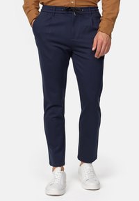 Pantalones de chándal azul marino con cintura elástica y cordón, pernera ajustada y dos bolsillos frontales. Combinados con zapatillas blancas y una camisa de botones marrón.