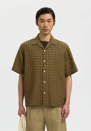 Homme portant une chemise marron texturée à manches courtes avec boutons par-dessus un sous-vêtement sombre, tenant un sac cabas beige sur un fond uni.