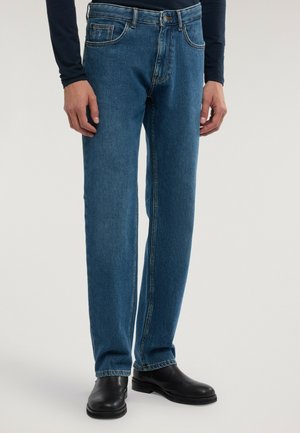 Man klädd i raka blå denimjeans, svarta läderskor och en marinblå tröja med långa ärmar står framför en enfärgad bakgrund.