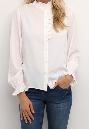 Hvid bluse med lange ærmer, flæsekant ved krave og manchetter, knappet foran, båret med blå jeans, den ene hånd i lommen og den anden afslappet.