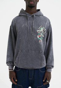 Grå hættetrøje lavet af blødt stof med en frontlomme, der har et blomstermotiv med en sort rose og tekst på venstre side.