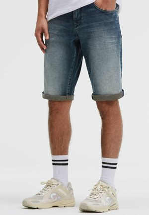 Mann trägt hochgekrempelte blaue Jeansshorts, weiße Sportsocken mit schwarzen Streifen und beige Sneakers, steht mit einer Hand in der Tasche.