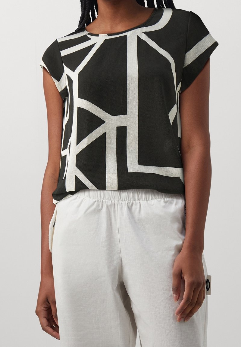 Femme portant un haut noir à manches courtes avec des lignes géométriques blanches, associé à un pantalon blanc ample avec une taille élastique.