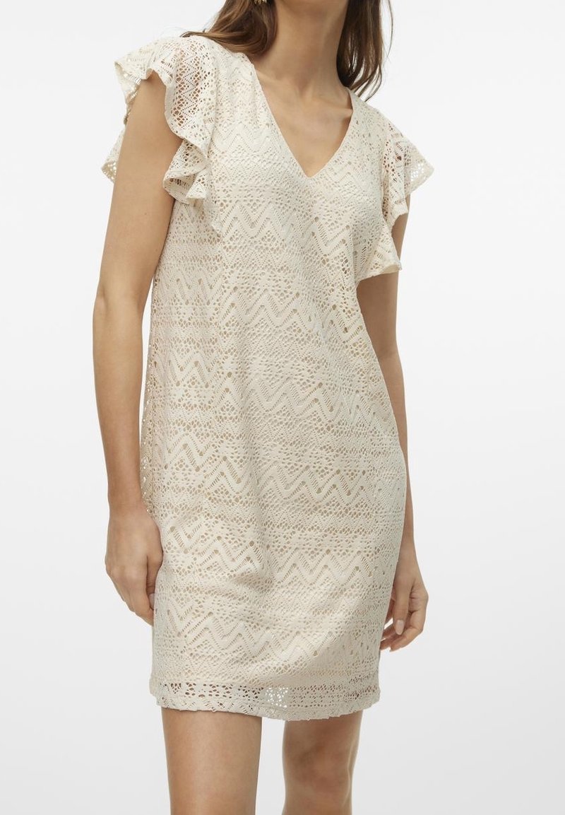 Femme portant une robe en dentelle crème avec des manches courtes à volants et un décolleté en V, debout devant un fond blanc uni.