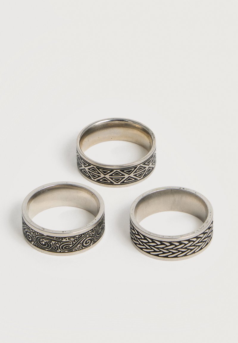 Tres anillos de plata con patrones grabados en negro de formas geométricas, remolinos y trenzados, dispuestos sobre una superficie blanca.