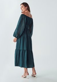 Tirkizna haljina s dugim prozirnim rukavima i slojevitom suknjom koja se nosi s jednim ramenom. Lagani materijal s naboranom teksturom, bijele sandale s petom.