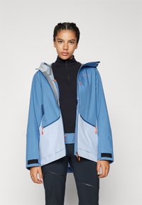 Chaqueta impermeable azul con detalles en gris y cremalleras naranjas, con capucha y grandes bolsillos frontales; tejido ligero y ajuste ajustable.