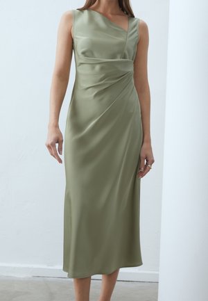 Femme portant une robe midi en satin vert olive clair sans manches avec un détail drapé asymétrique à la taille sur un fond uni.