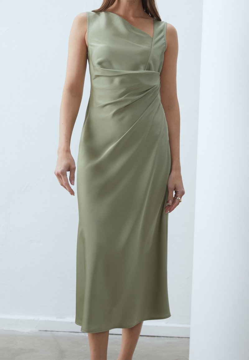 Femme portant une robe midi en satin vert olive clair sans manches avec un détail drapé asymétrique à la taille sur un fond uni.