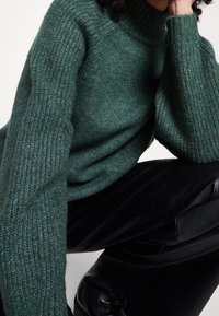 Personne portant un pull vert en maille épaisse et un pantalon en cuir noir, assise avec le coude appuyé sur le genou, sur un fond blanc.