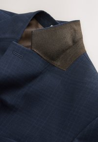 Blue suit jacket with a brown contrasting inner collar, showing detailed stitching and a subtle check pattern on the fabric.