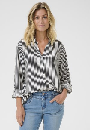 Striped button-up shirt in black and white, featuring a collar, rolled sleeves, and a loose fit. Paired with light blue jeans.