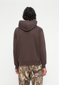 Persona de espaldas vistiendo una sudadera marrón oscura simple y pantalones de camuflaje contra un fondo blanco.