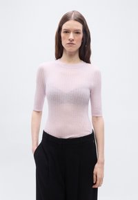 Νεαρή γυναίκα με μ ακριά καστανά μαλλιά φορά ένα ανοιχτό ροζ ριμπ μπλουζάκι με κοντά μανίκια και άνετο μαύρο παντελόνι, στέκεται μπροστά σε απλό φόντο.