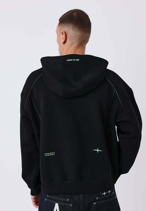 Homme vu de dos portant un sweat à capuche noir avec le texte « CHANGE THE GAME » sur la capuche et « PROJECT X PARIS » près du bas, associé à un jean sombre.