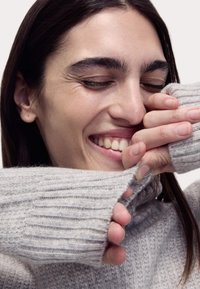Maglione a coste grigio chiaro con colletto alto, caratterizzato da un tessuto testurizzato e maniche aderenti, tirato delicatamente da mani.