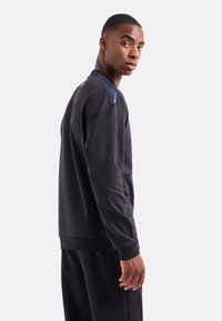 Sudadera de color gris oscuro con un acento texturizado en los hombros, escote redondeado y puños acanalados, combinada con pantalones a juego, modelo de perfil.