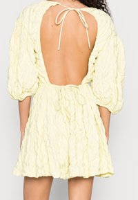 Gelbes Kleid mit Puffärmeln und strukturiertem, gekräuseltem Design. Verfügt über einen offenen Rücken, der am Nacken und in der Taille gebunden ist, um eine verstellbare Passform zu gewährleisten.