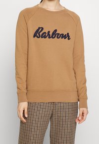 Brown sweatshirt with navy "Barbour" embroidery, raglan sleeves, ribbed cuffs, and hem. Worn over patterned trousers; smooth fabric texture.