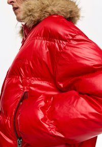 Czerwona puchowa kurtka z połyskującym wykończeniem, wyposażona w kaptur obszyty futrem, kieszenie na suwak oraz wytłoczone logo na rękawie.