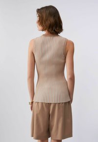 Femme aux cheveux bruns courts portant un haut sans manches beige et blanc à rayures et un short large beige, debout devant un fond clair uni.