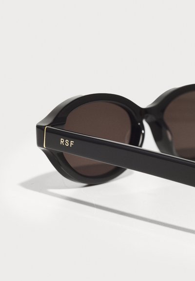 Lunettes de soleil noires avec des verres ronds et sombres au fini brillant. La monture a une texture lisse et un accent doré portant l'inscription "RSF" sur la branche.