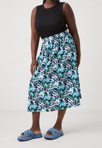 Mustat hihattomat topit yhdistettynä pitkään, moniväriseen hameeseen, jossa on sinisiä, vihreitä ja mustia abstrakteja kuvioita; siniset ristikkäisandaalit.