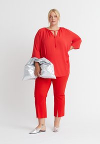 Rote Bluse mit einem Schlüssellochdetail, kombiniert mit roten 3/4-Hosen mit seitlichen Schlitzen. Hält eine silberne, reflektierende Jacke. Silberne Slipper.