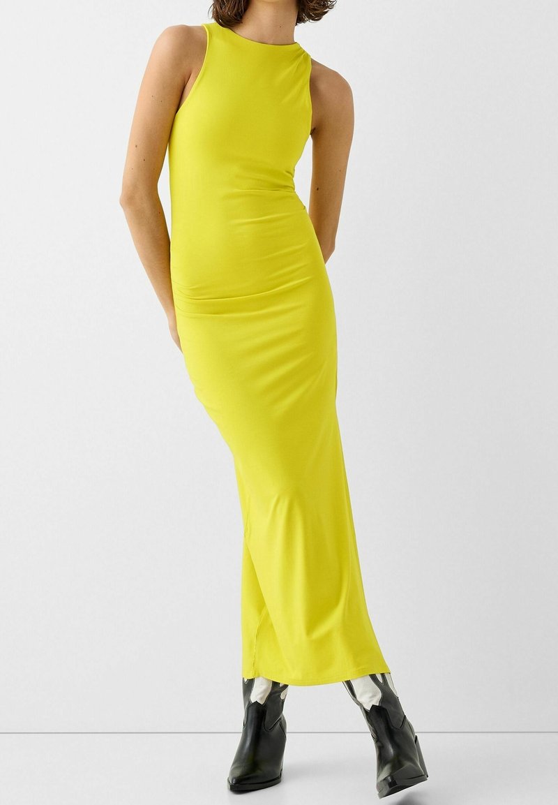 Bezramenné, těsně přiléhající žluté maxi šaty s kulatým výstřihem, vyrobené z hladkého materiálu. Nosené s černými kotníkovými botami pro kontrast.