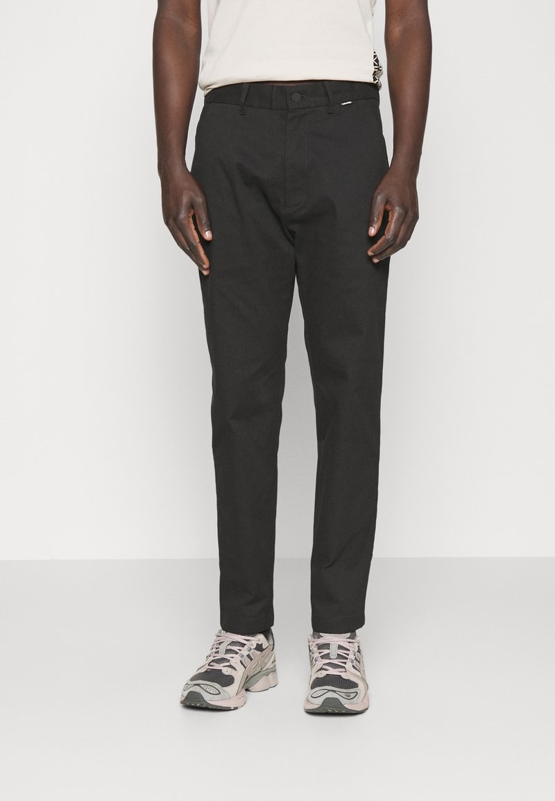 Homme portant un pantalon noir slim et des baskets de sport blanches, grises et roses, debout devant un fond clair uni.