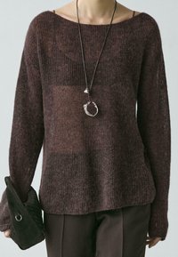 Bruine, losgebreide trui met een brede halslijn en lange mouwen. Draagt donkere broek; heeft een zilveren hanger en een gestructureerde zwarte tas.