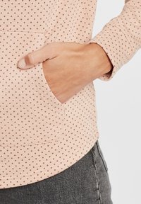 Maglietta a maniche lunghe rosa chiaro con un motivo a pois rosso scuro, dotata di tasca frontale e vestibilità rilassata. Tessuto morbido e polsini a costine.